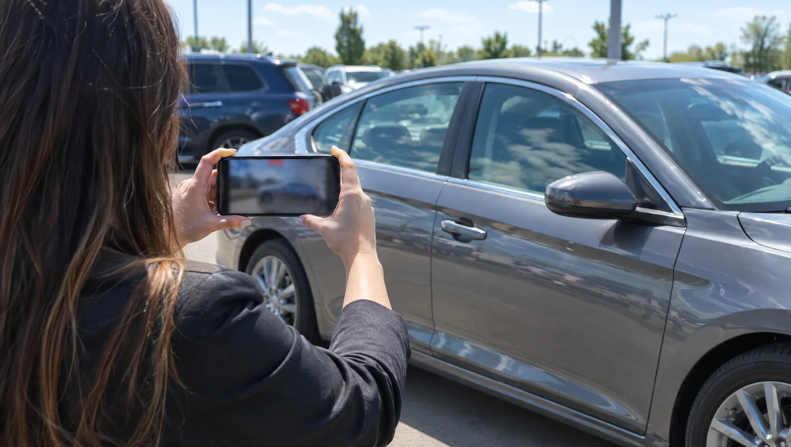 Salesperson holding a smartphone to film the side of a gray sedan on a sunny dealership lot